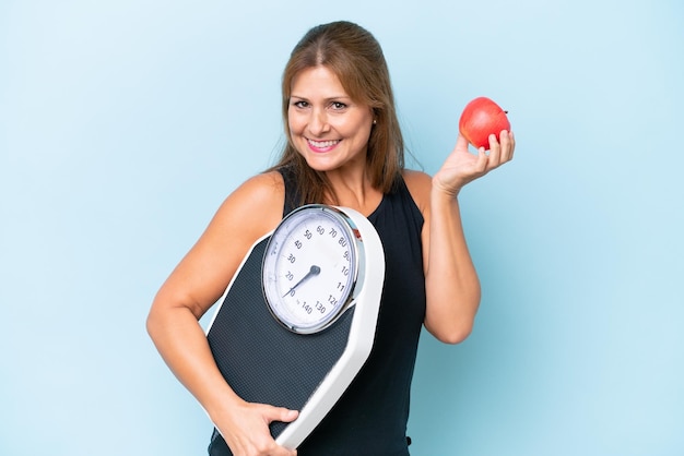 Mujer caucásica de mediana edad aislada de fondo azul con máquina de pesaje y con una manzana