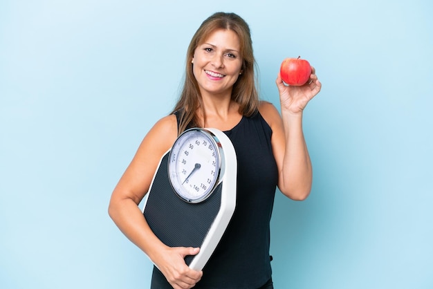 Mujer caucásica de mediana edad aislada de fondo azul con máquina de pesaje y con una manzana