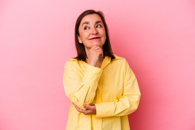 Mujer caucásica de mediana edad aislada en un fondo rosado pensando y mirando hacia arriba, siendo reflexiva, contemplando, teniendo una fantasía.