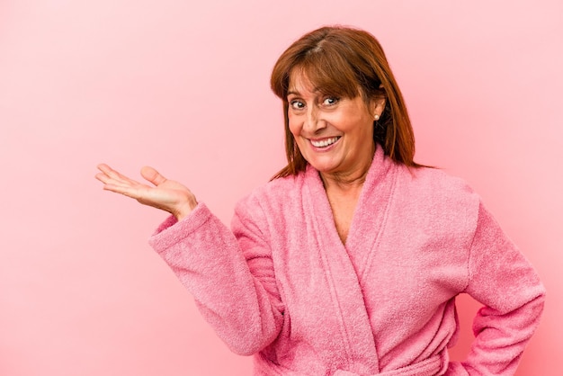 Mujer caucásica de mediana edad con una bata de baño aislada sobre fondo rosa que muestra un espacio de copia en una palma y sosteniendo otra mano en la cintura.