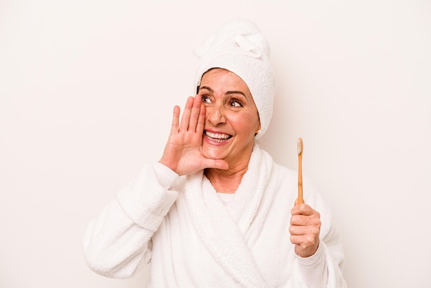 Mujer caucásica de mediana edad con una bata de baño sosteniendo un cepillo de dientes aislado de fondo blanco gritando y sosteniendo la palma cerca de la boca abierta