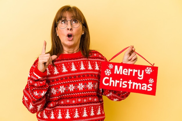 Mujer caucásica de mediana edad con cartel de 'Feliz Navidad' aislado sobre fondo amarillo apuntando hacia arriba con la boca abierta.