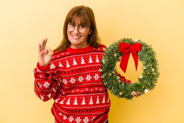Mujer caucásica de mediana edad celebrando la Navidad con corona aislado sobre fondo amarillo alegre y confiado mostrando gesto ok.