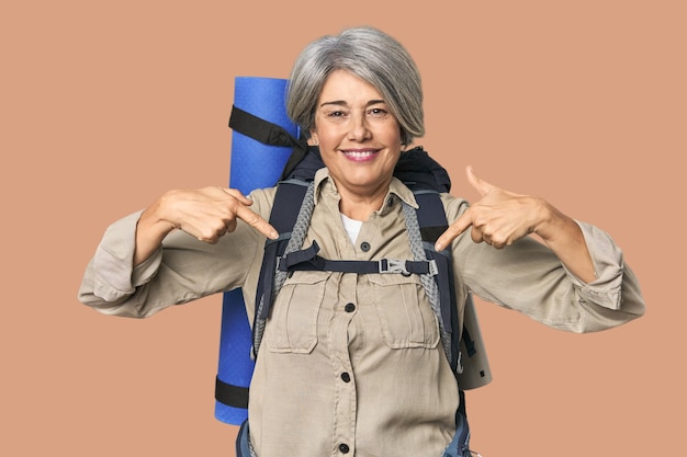 Mujer caucásica de mediana edad con equipo de senderismo sorprendida señalando con el dedo sonriendo ampliamente