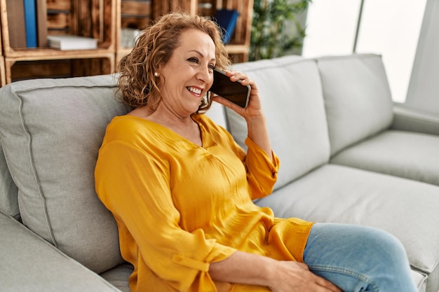 Mujer caucásica de mediana edad hablando por teléfono inteligente sentada en el sofá de casa.