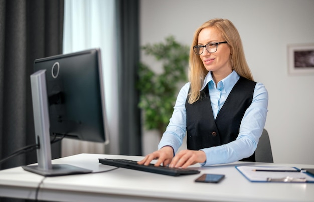 Mujer caucásica de mediana edad con ropa formal y anteojos escribiendo en una computadora moderna mientras está sentada en la oficina doméstica Concepto de freelance y tecnología