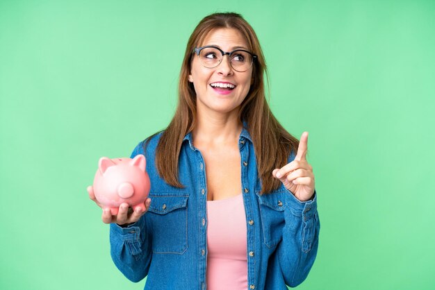 Mujer caucásica de mediana edad sosteniendo una alcancía sobre un fondo aislado con la intención de darse cuenta de la solución mientras levanta un dedo