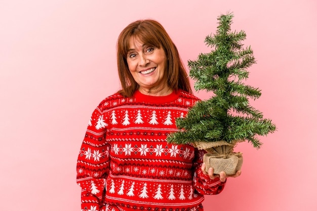 Mujer caucásica de mediana edad sosteniendo el árbol de Navidad aislado sobre fondo rosa feliz, sonriente y alegre.