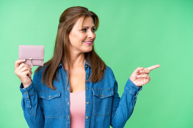 Mujer caucásica de mediana edad sosteniendo una billetera sobre un fondo aislado apuntando hacia un lado para presentar un producto