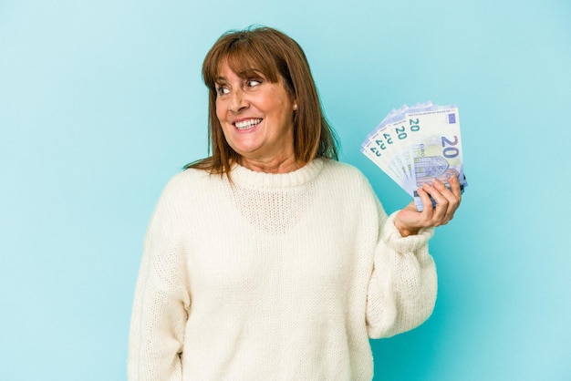 Mujer caucásica de mediana edad sosteniendo billetes de banco aislados sobre fondo azul se ve a un lado sonriente, alegre y agradable.