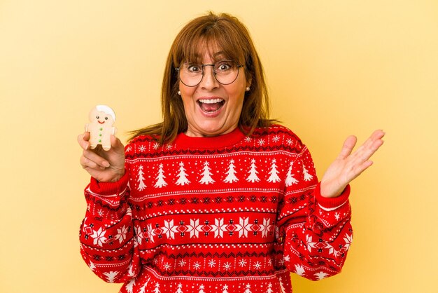 Mujer caucásica de mediana edad sosteniendo una galleta de Navidad aislada sobre fondo amarillo recibiendo una agradable sorpresa, emocionada y levantando las manos.