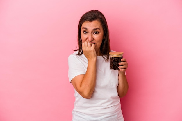 Mujer caucásica de mediana edad sosteniendo una jarra de café aislada en un fondo rosado mordiéndose las uñas, nerviosa y muy ansiosa.