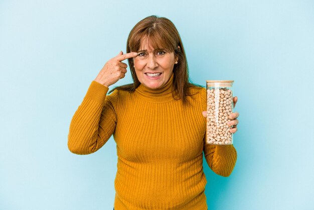 Mujer caucásica de mediana edad sosteniendo un tarro de garbanzos aislado sobre fondo azul que muestra un gesto de decepción con el dedo índice.