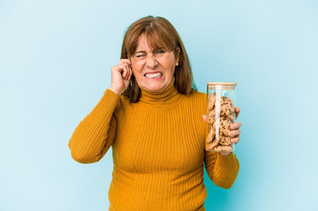 Mujer caucásica de mediana edad con tarro de galletas aislado sobre fondo azul cubriendo los oídos con las manos.