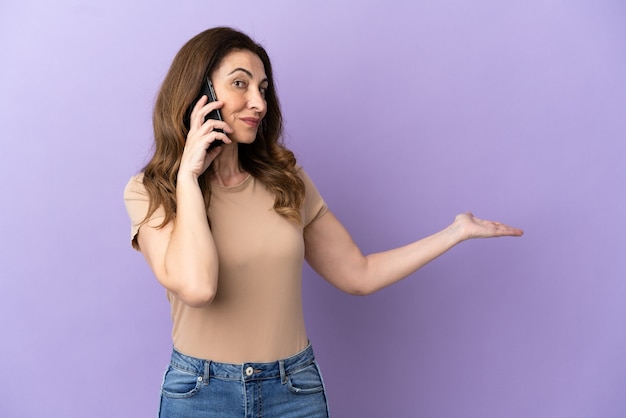 Mujer caucásica de mediana edad con teléfono móvil aislado sobre fondo púrpura extendiendo las manos hacia el lado para invitar a venir