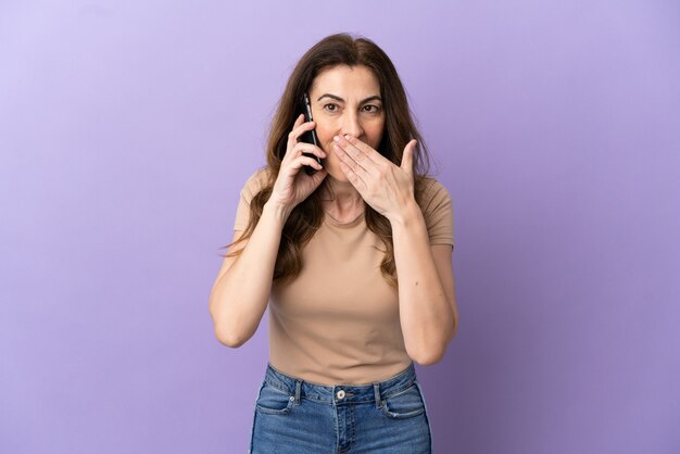 Mujer caucásica de mediana edad con teléfono móvil aislado sobre fondo púrpura feliz y sonriente cubriendo la boca con la mano