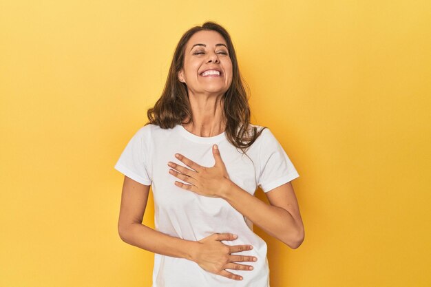 Mujer caucásica de mediana edad vestida de amarillo se ríe alegremente y se divierte manteniendo las manos en el estómago