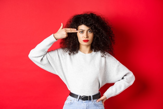 Mujer caucásica molesta con el pelo rizado, mostrando el signo de pistola de mano en la cabeza, volar su mente, de pie molesta contra el fondo rojo.