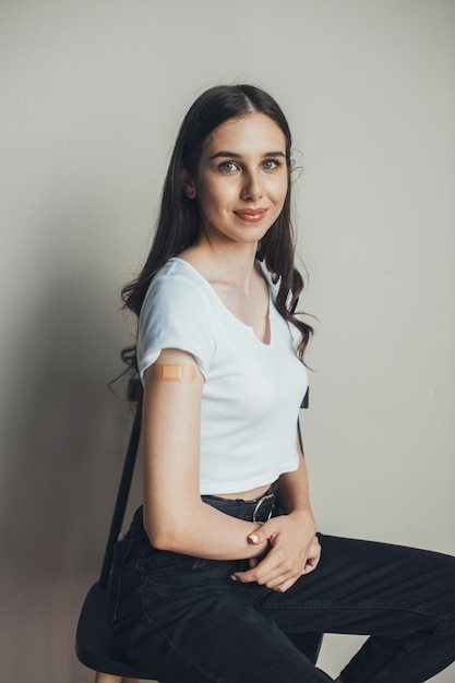 Mujer caucásica mostrando el brazo vacunado después de la inyección sobre fondo blanco sentado en una silla y mirar ...