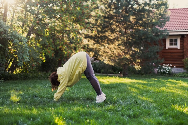 Mujer caucásica obesa haciendo ejercicio al aire libre en ropa deportiva con los pies y las manos puestos en el suelo sobre hierba fresca en el patio trasero con una casa de campo de madera y árboles altos en el fondo Cuerpo positivo