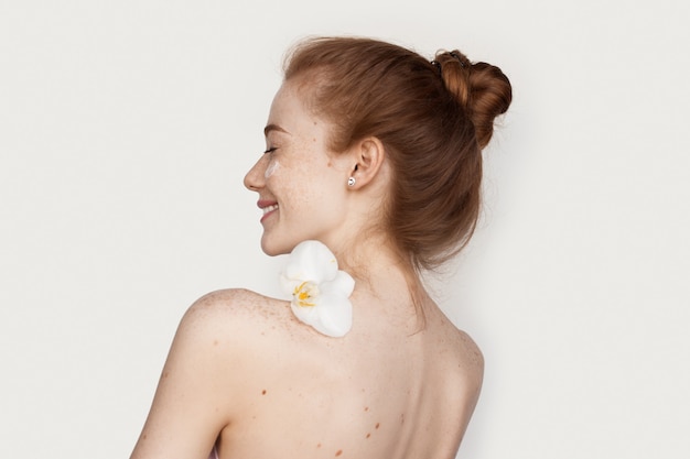 Mujer caucásica con pecas y cabello rojo sosteniendo una flor sobre sus hombros sonriendo con una crema en la cara en la pared blanca