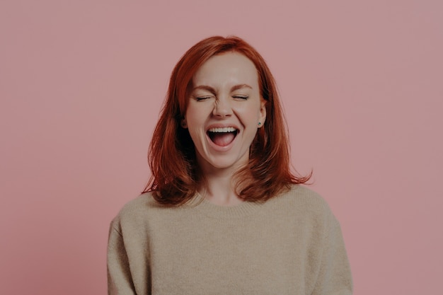 Foto mujer caucásica pelirroja llena de alegría riendo positivamente, con los ojos cerrados, expresando emociones positivas después de escuchar una broma hilarante o una anécdota, aislada sobre fondo rosa. concepto de expresiones faciales