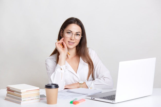 Mujer caucásica con el pelo largo, usa gafas, mira seminario web en la computadora portátil, prepara el proyecto, hace la tarea, bebe café para llevar, aislado en blanco