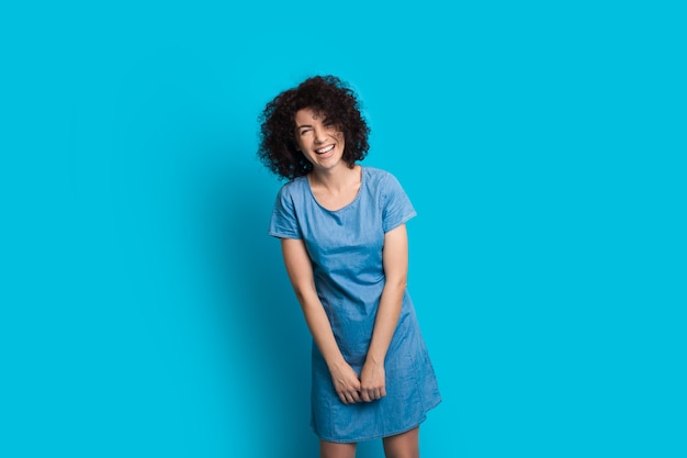 Mujer caucásica de pelo rizado posando con un bonito vestido sobre un fondo azul mientras sonríe alegremente