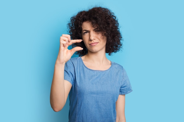 Mujer caucásica de pelo rizado posando sobre una pared azul y gesticulando el pequeño cartel