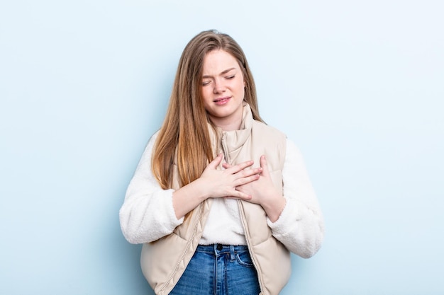 Mujer caucásica de pelo rojo que parece triste, herida y con el corazón roto sosteniendo ambas manos cerca del corazón llorando y sintiéndose deprimida