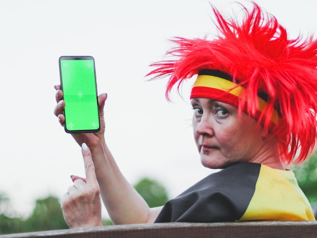 Mujer caucásica con peluca de bandera belga sosteniendo un smartphone con pantalla verde