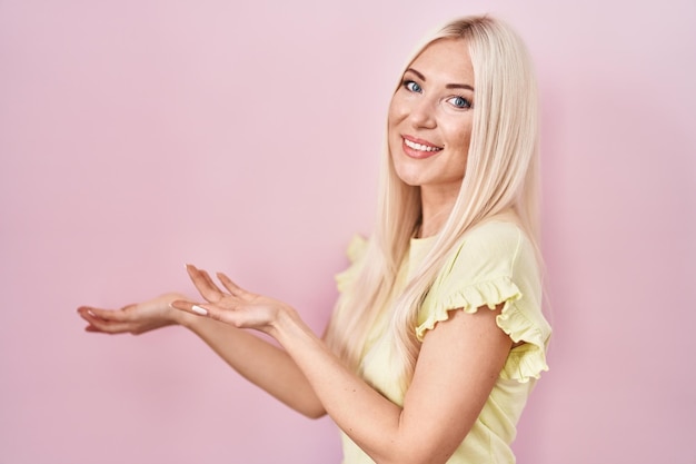 Mujer caucásica de pie sobre fondo rosa invitando a entrar sonriendo natural con la mano abierta