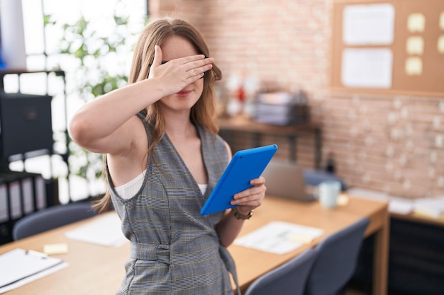 Mujer caucásica que trabaja en la oficina con gafas sonriendo y riendo con la mano en la cara cubriendo los ojos por sorpresa. concepto ciego.