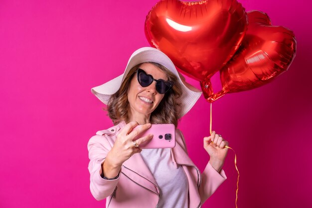 Una mujer caucásica rubia divirtiéndose con un sombrero blanco y gafas de sol en una discoteca con algunos globos de corazón tomándose una selfie con su teléfono móvil de fondo rosa