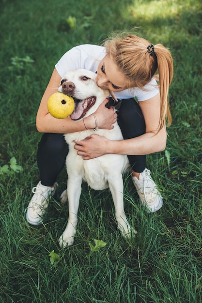 Mujer caucásica rubia está abrazando a su perro mientras juega en un parque verde con una pelota