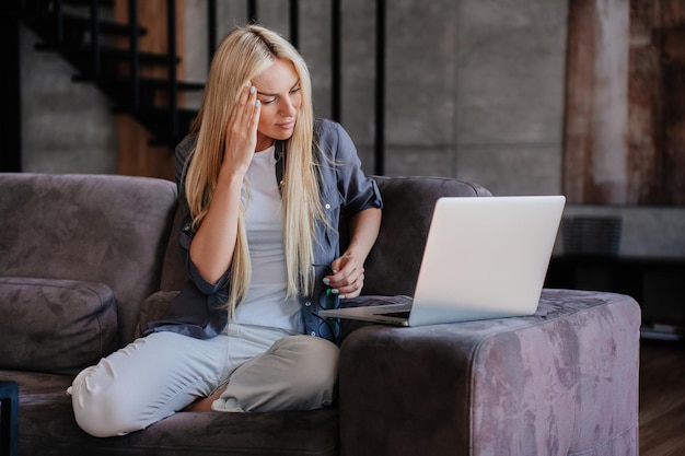 Mujer caucásica rubia exhausta con camisa gris y pantalones blancos sentada en un sofá con una laptop tocando