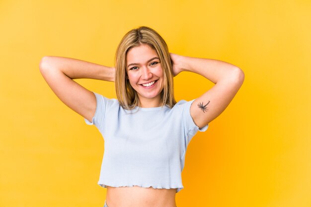 Mujer caucásica rubia joven aislada en un espacio amarillo que se siente confiada, con las manos detrás de la cabeza.