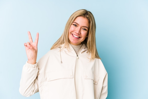 La mujer caucásica rubia joven aislada mostrando la muestra de la victoria y sonriendo ampliamente.