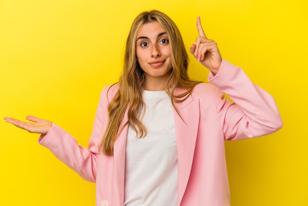 Mujer caucásica rubia joven aislada en la pared amarilla que sostiene y que muestra un producto en la mano.