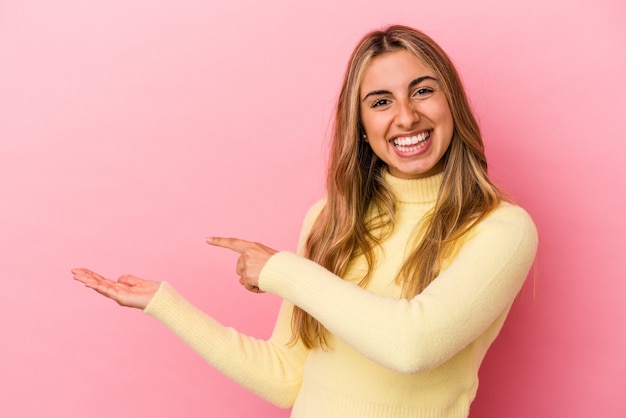 Mujer caucásica rubia joven aislada en la pared rosada emocionada sosteniendo un espacio de la copia en la palma.