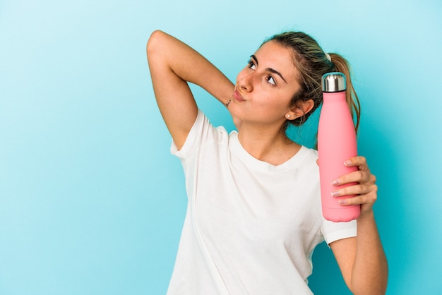 Mujer caucásica rubia joven que sostiene un termo que toca la parte posterior de la cabeza, pensando y haciendo una elección.