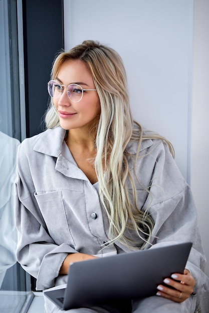 Mujer caucásica rubia en ropa doméstica casual usando computadora portátil y sentada en el alféizar de la ventana...