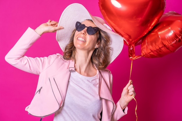 Una mujer caucásica rubia con un sombrero blanco y gafas de sol en una discoteca con algunos globos de corazón tomando un selfie de fondo rosa