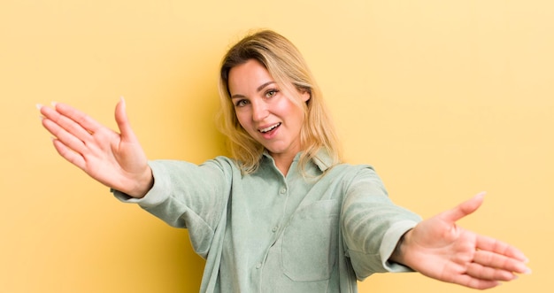 Mujer caucásica rubia sonriendo alegremente dando un cálido abrazo cariñoso de bienvenida sintiéndose feliz y adorable