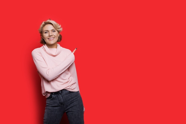 Mujer caucásica rubia sonriendo y apuntando a la pared roja con espacio libre cerca de ella con un suéter rosa