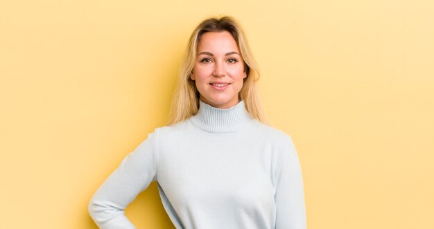 Mujer caucásica rubia sonriendo felizmente con una mano en la cadera y confiada actitud positiva, orgullosa y amistosa