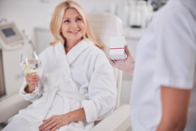 Mujer caucásica rubia con vaso de agua en la mano escuchando a su médico en el gabinete