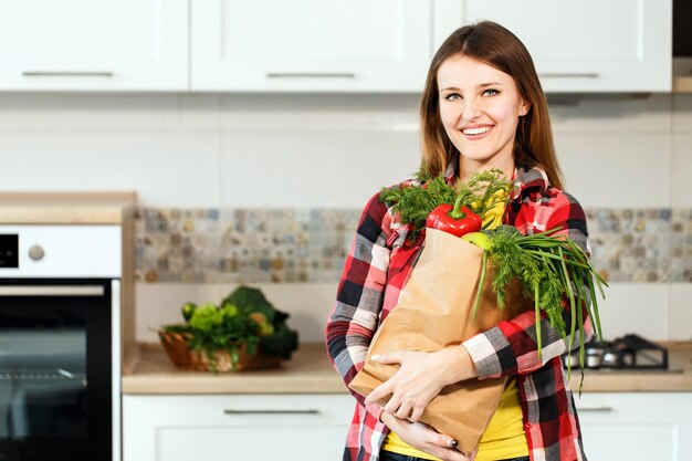 Mujer caucásica satisfecha con camisa casual a cuadros sosteniendo un paquete de compras con alimentos orgánicos frescos de pie felizmente en medio de una espaciosa cocina blanca