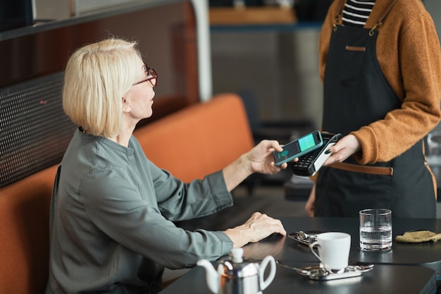 Mujer caucásica senior de pelo rubio sentado a la mesa en la cafetería y poner el teléfono inteligente en el terminal de pago en manos de la camarera en el delantal