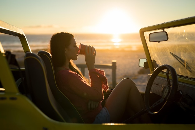 Mujer caucásica sentada en un buggy de playa junto al mar durante la bebida al atardecer. descanso en la playa en un viaje por carretera de vacaciones de verano.